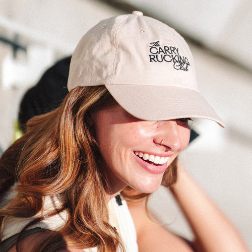 Woman wearing a beige cap with 'The Carry Rucking Club' text, smiling outdoors.