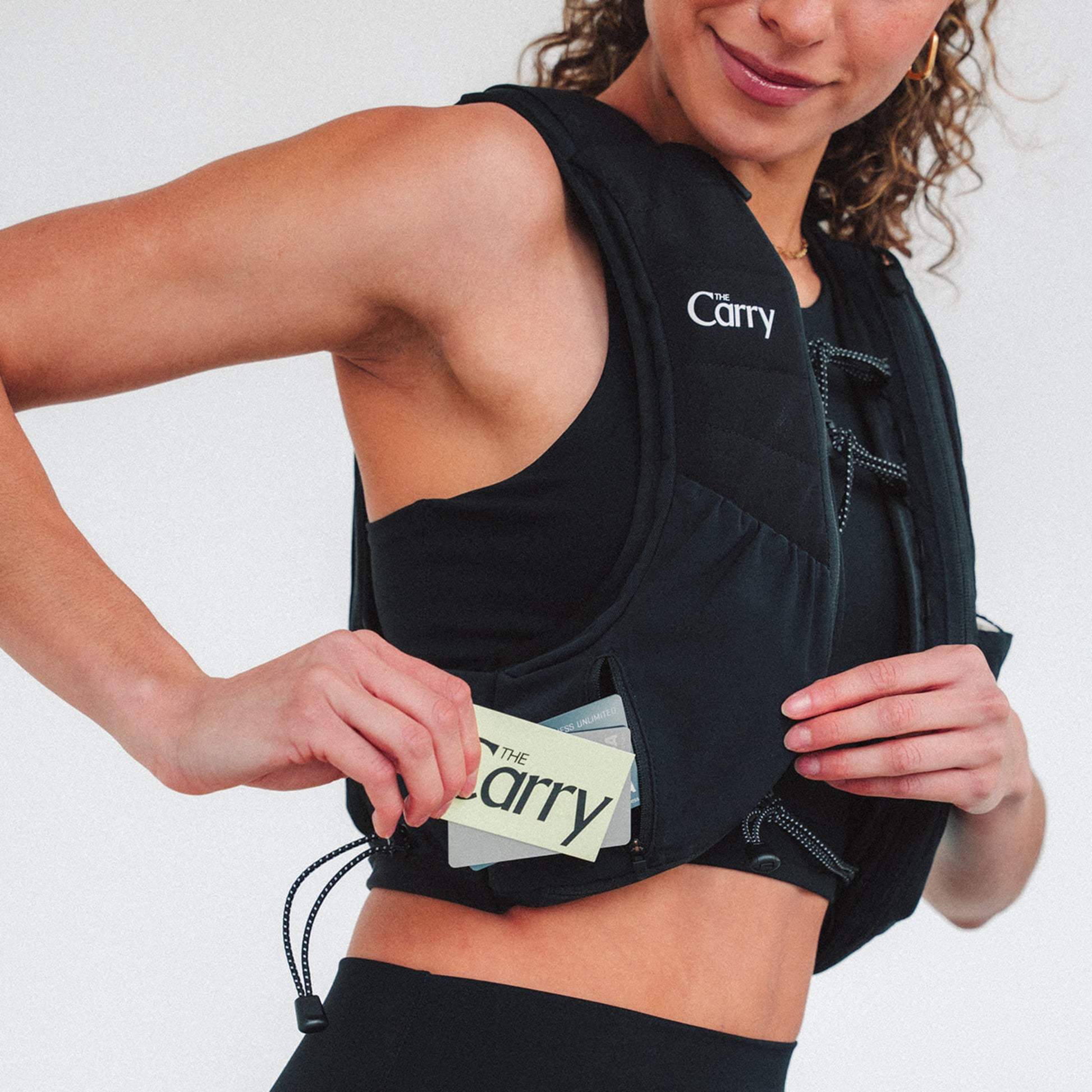 Woman wearing The Carry weighted vest in black against a plain background, pulling out cards from shell pocket