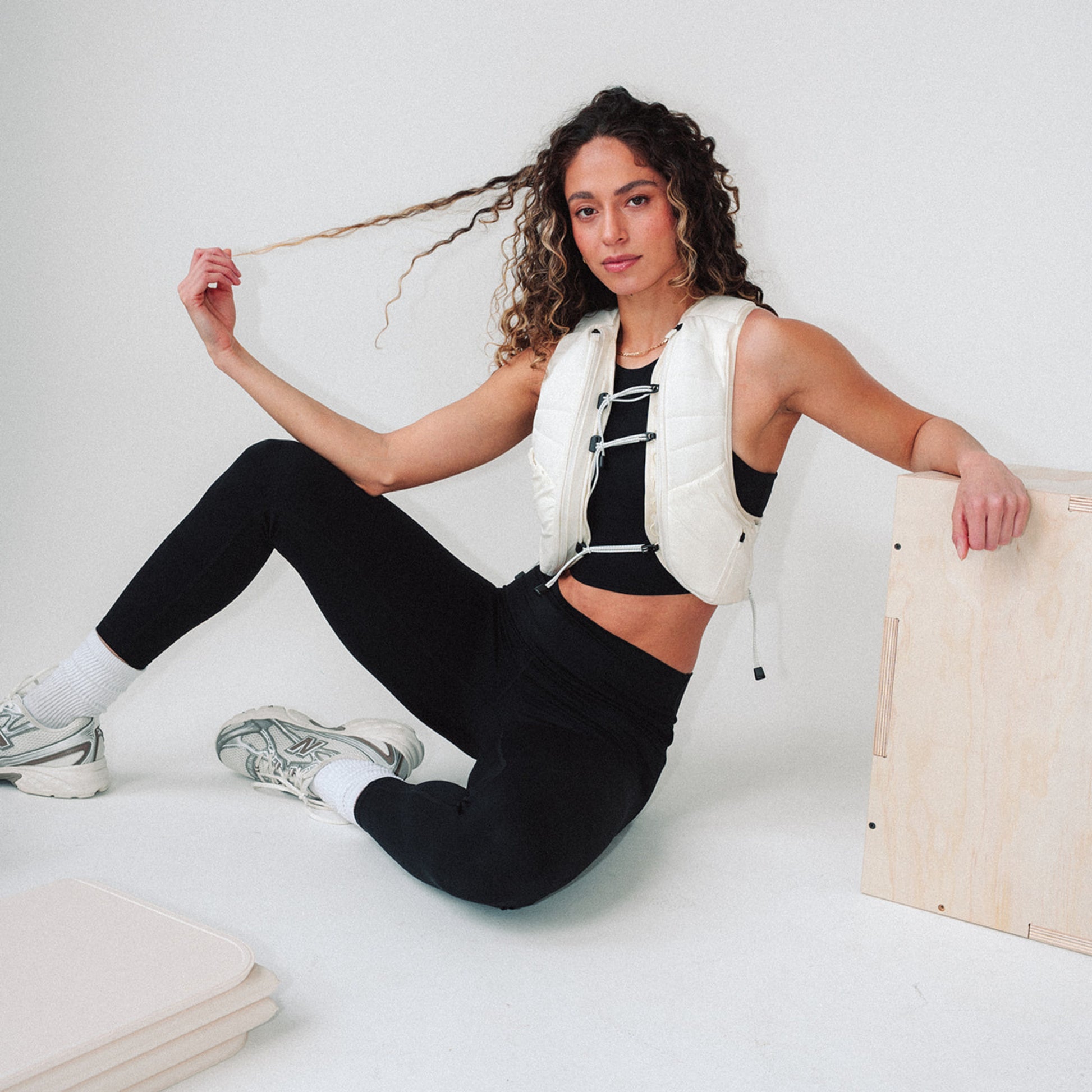 Woman wearing The Carry weighted vest in cream, sitting on the floor next to a plyo box and yoga mat against a plain background