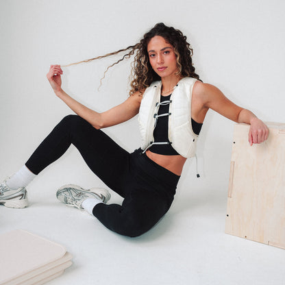 Woman wearing The Carry weighted vest in cream, sitting on the floor next to a plyo box and yoga mat against a plain background